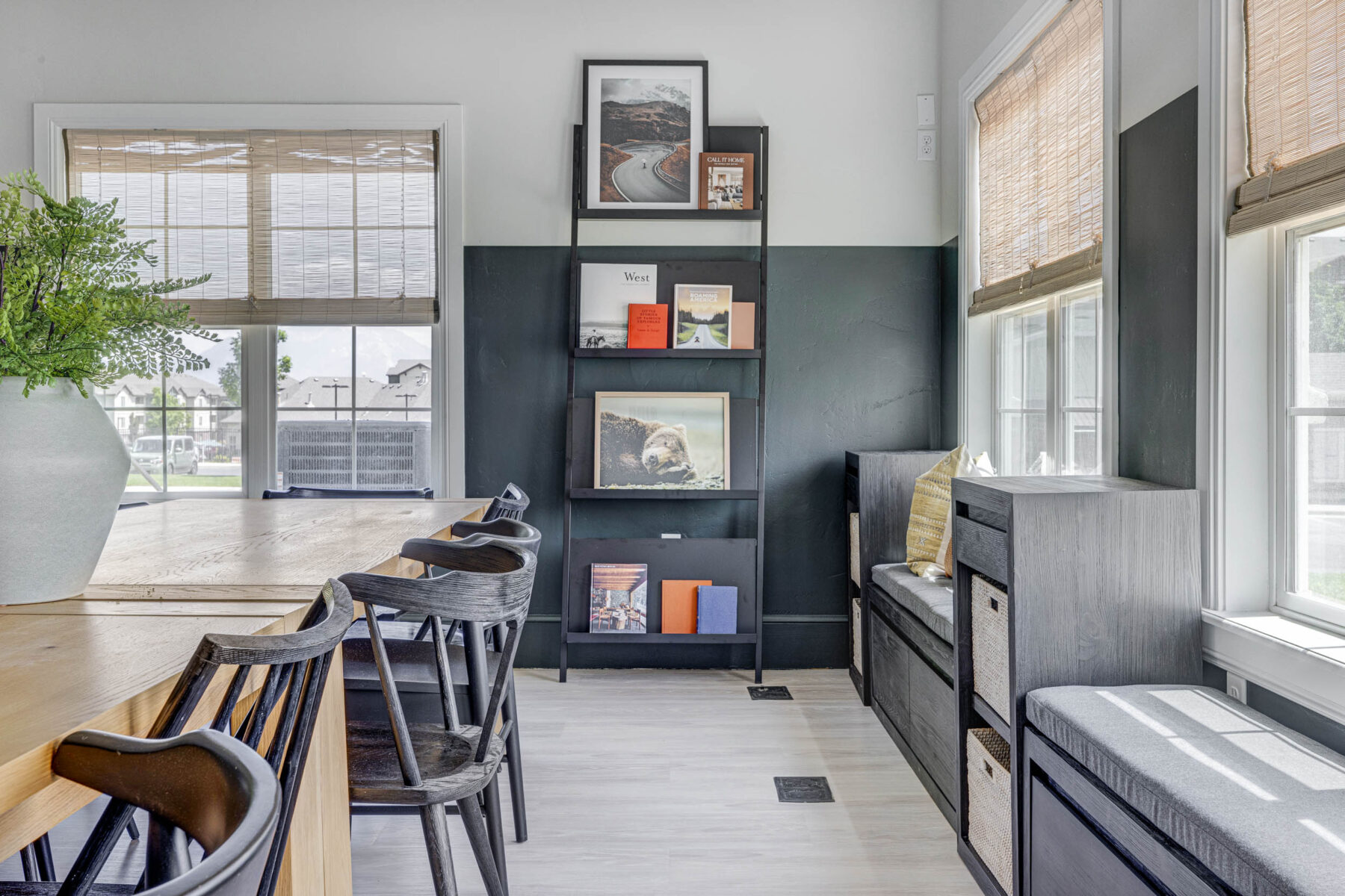 Modern dining area in apartments in Herriman UT features black chairs around a wooden table, a vase with green foliage, tall shelving with books and art, cozy bench seating, and large windows with woven blinds letting in natural light.