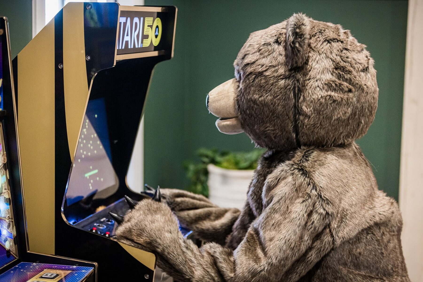 A person wearing a bear costume plays a vintage arcade game machine in a room with green walls and a potted plant in the background.