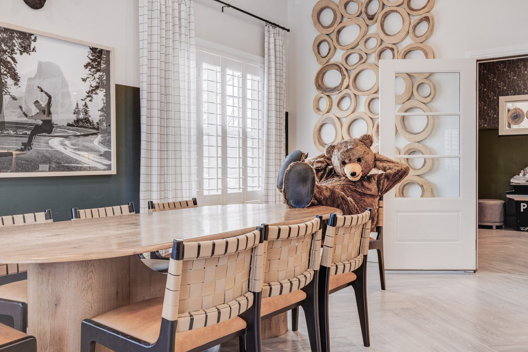 A large teddy bear lounges in an armchair at the head of a long wooden dining table surrounded by woven chairs, giving these apartments in Herriman UT a cozy, whimsical touch. Art and round wall decor hang on light walls as sunlight streams through the windows.