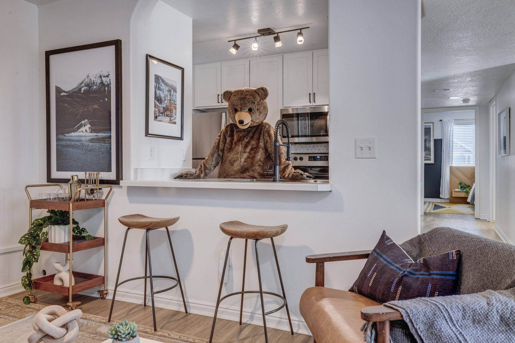 A person wearing a bear costume stands behind a modern kitchen counter in a cozy, stylish apartment. There are two bar stools, framed photos, and a small bar cart—perfect touches for unique apartments in Herriman UT.