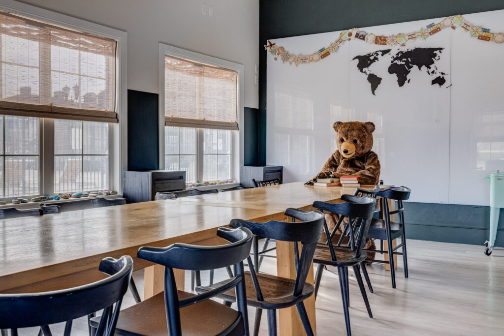 A large teddy bear sits at a long wooden table in a classroom with black chairs, a whiteboard featuring a world map, and windows with bamboo blinds.