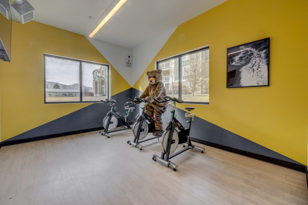 A person in a bear costume sits on a stationary bike in a brightly lit gym room with yellow and gray walls, two windows, and a framed photo on the wall.