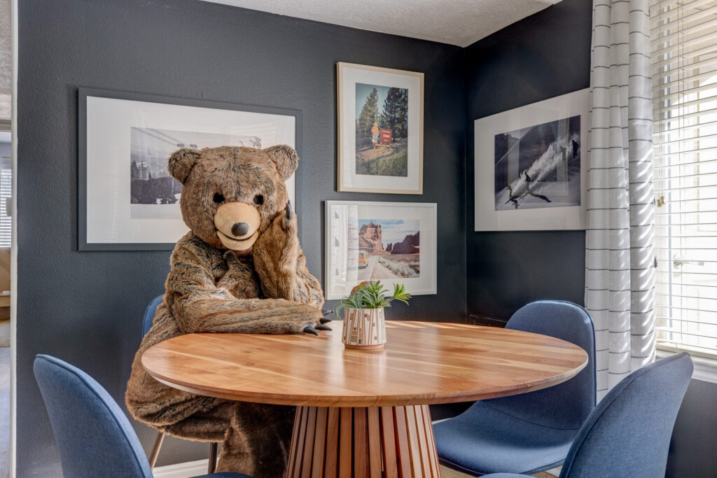 A person in a bear costume sits at a round wooden table in a modern dining area with blue chairs, plant centerpiece, and framed photos on dark walls near a window with striped curtains.