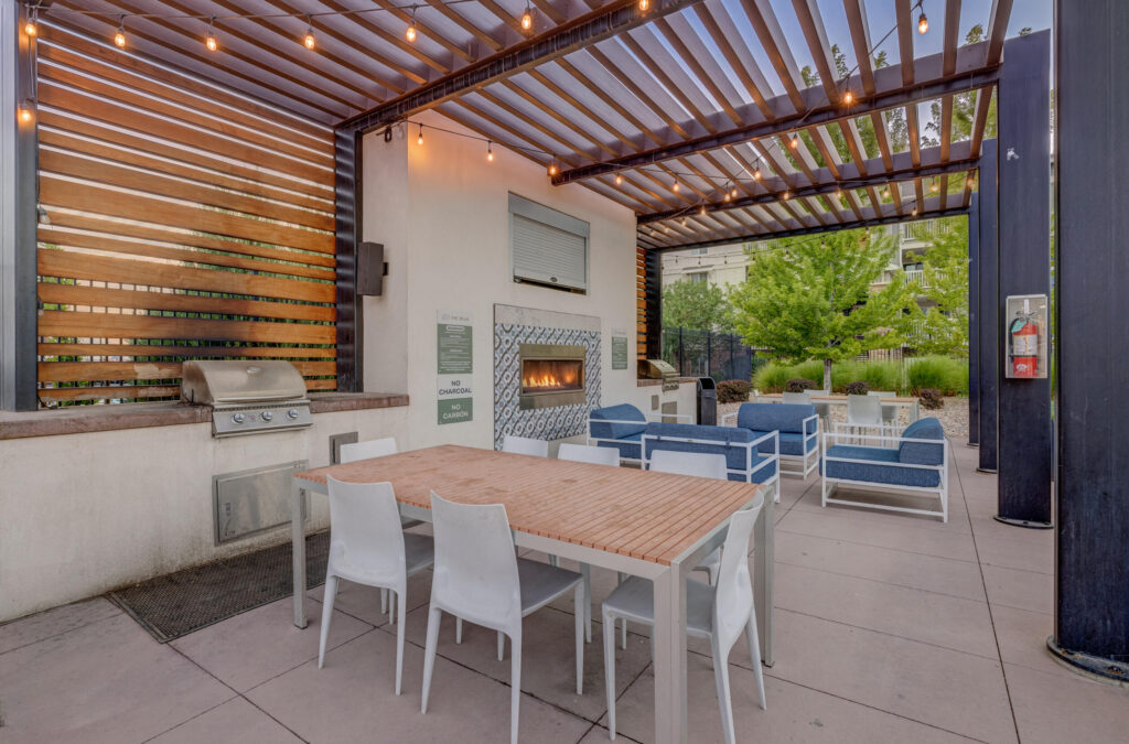 Modern outdoor patio with a pergola, string lights, grill, fireplace, wooden dining table with white chairs, and blue cushioned lounge seating, surrounded by greenery and a nearby building.