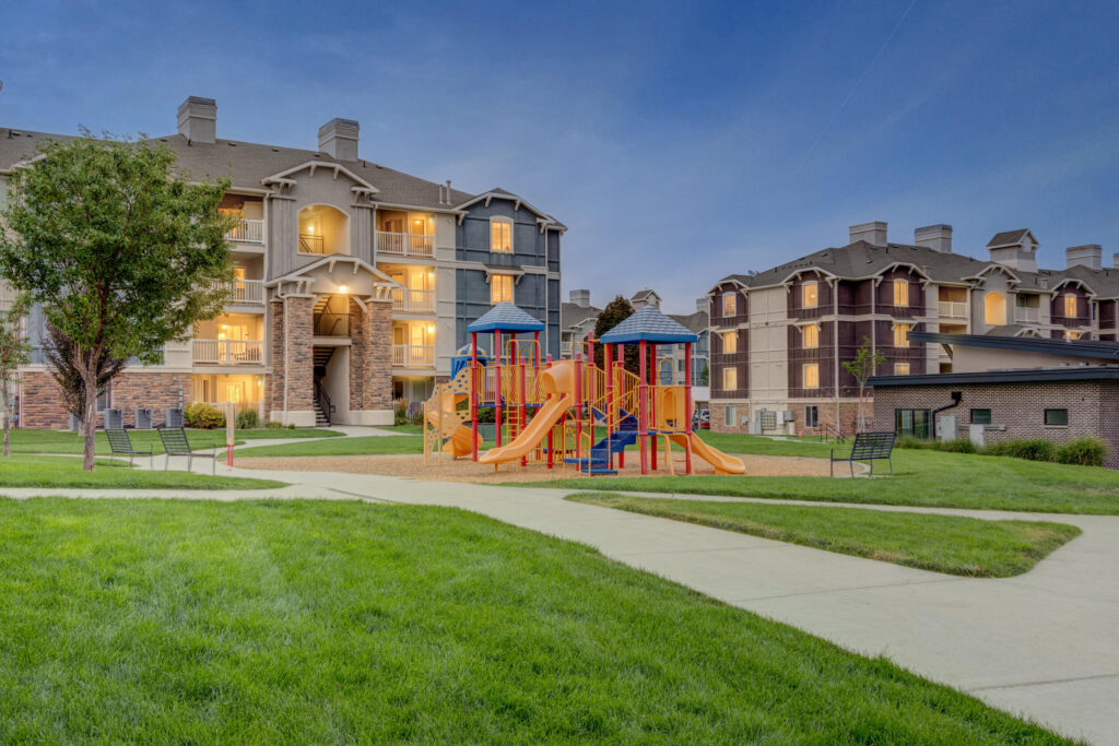 Colorful playground with slides and climbing structures sits on sand, surrounded by green grass. Two multi-story apartment buildings with lit windows are in the background during early evening.