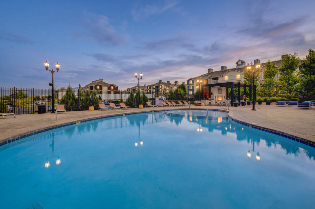 Outdoor swimming pool with clear blue water, surrounded by lounge chairs, lampposts, and greenery. Apartment buildings and a clubhouse are visible in the background under a dusk sky.