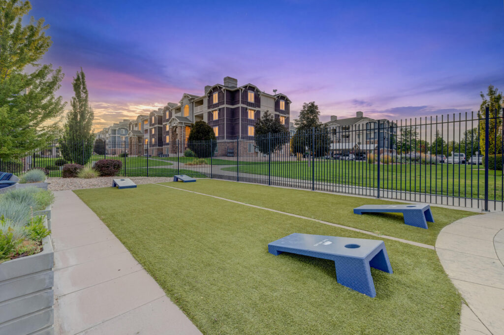 A fenced outdoor area with artificial grass, cornhole boards, and apartment buildings in the background under a vibrant sunset sky.