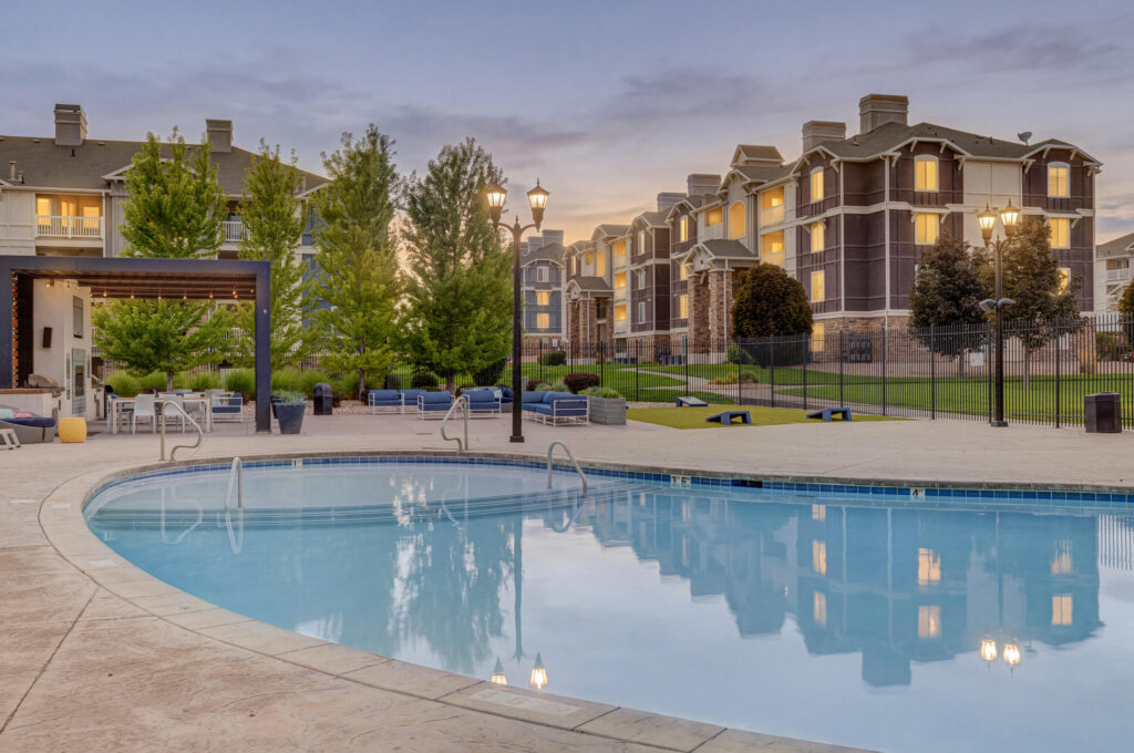 A curved outdoor swimming pool with poolside seating, surrounded by trees and apartment buildings at sunset, soft lights glowing from the windows.