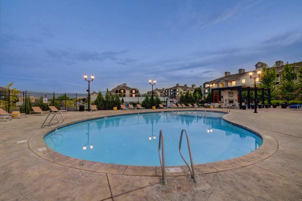 A round outdoor swimming pool with metal handrails, surrounded by lounge chairs and lampposts, in a courtyard with apartment buildings and trees at dusk. The sky is partly cloudy.